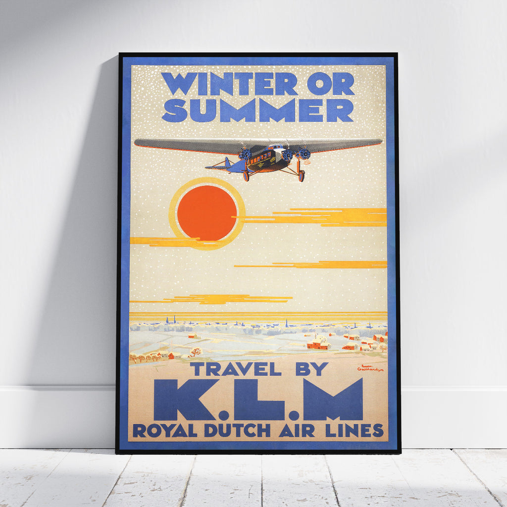A framed vintage-style travel poster leaning against a white wall on a light floor. The poster reads “WINTER OR SUMMER” and “TRAVEL BY K.