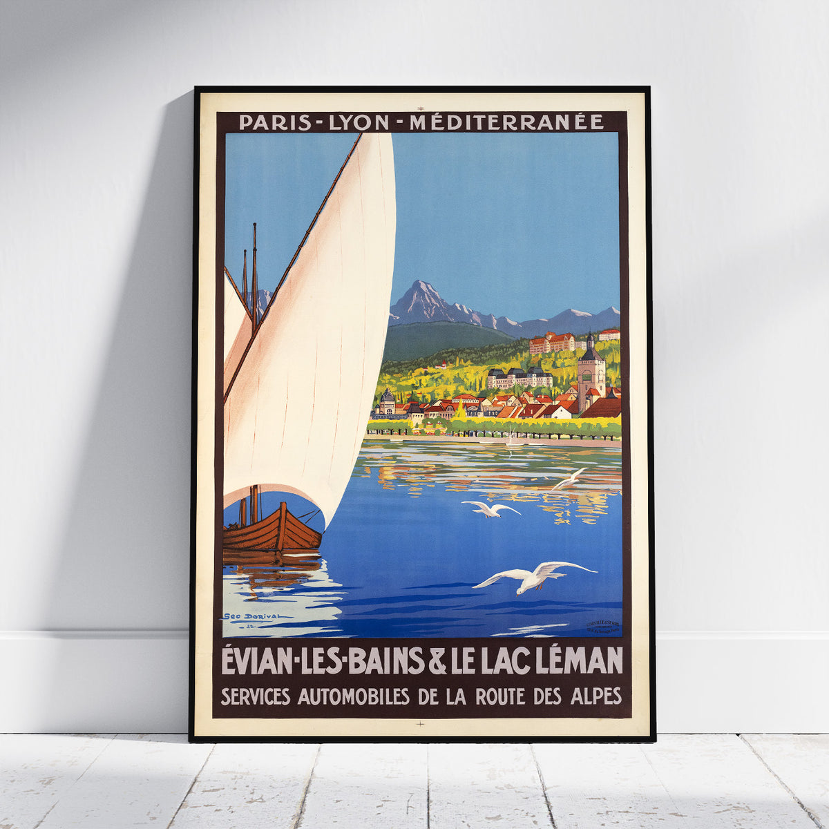 A framed vintage travel poster leaning against a white wall on a light floor. The poster features a sailboat on blue water with mountains and a town in the background, and text reading 'PARIS - LYON - MÉDITERRANÉE' and 'ÉVIAN-LES-BAINS & LE LAC LÉMAN'.