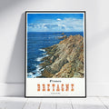A framed vintage-style travel poster of Brittany, France, leaning against a white wall on a light wood floor. The poster features a scenic photograph of a rocky coastline and blue ocean under a cloudy sky, with 'France BRETAGNE' printed in orange and black text on a white background at the bottom.