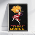 A framed vintage-style poster leaning against a white wall on a light floor. The poster shows a woman in a flowing red dress with a large yellow hair swirl holding a glass, on a black background, with the text 'COGNAC MONNET' and the phrase '.