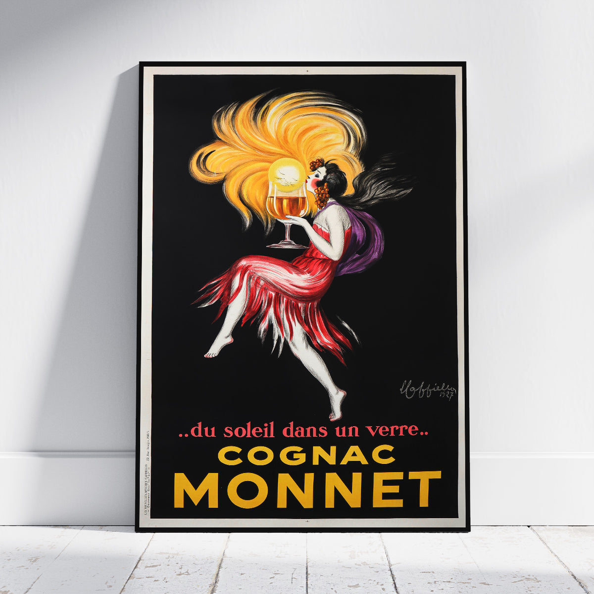 A framed vintage-style poster leaning against a white wall on a light floor. The poster shows a woman in a flowing red dress with a large yellow hair swirl holding a glass, on a black background, with the text 'COGNAC MONNET' and the phrase '.