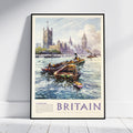 A framed vintage-style travel poster of London, Britain, leaning against a white wall on a light wood floor. The artwork shows boats on the River Thames with the Houses of Parliament and Big Ben in the background, and the word 'BRITAIN' printed prominently at the bottom.