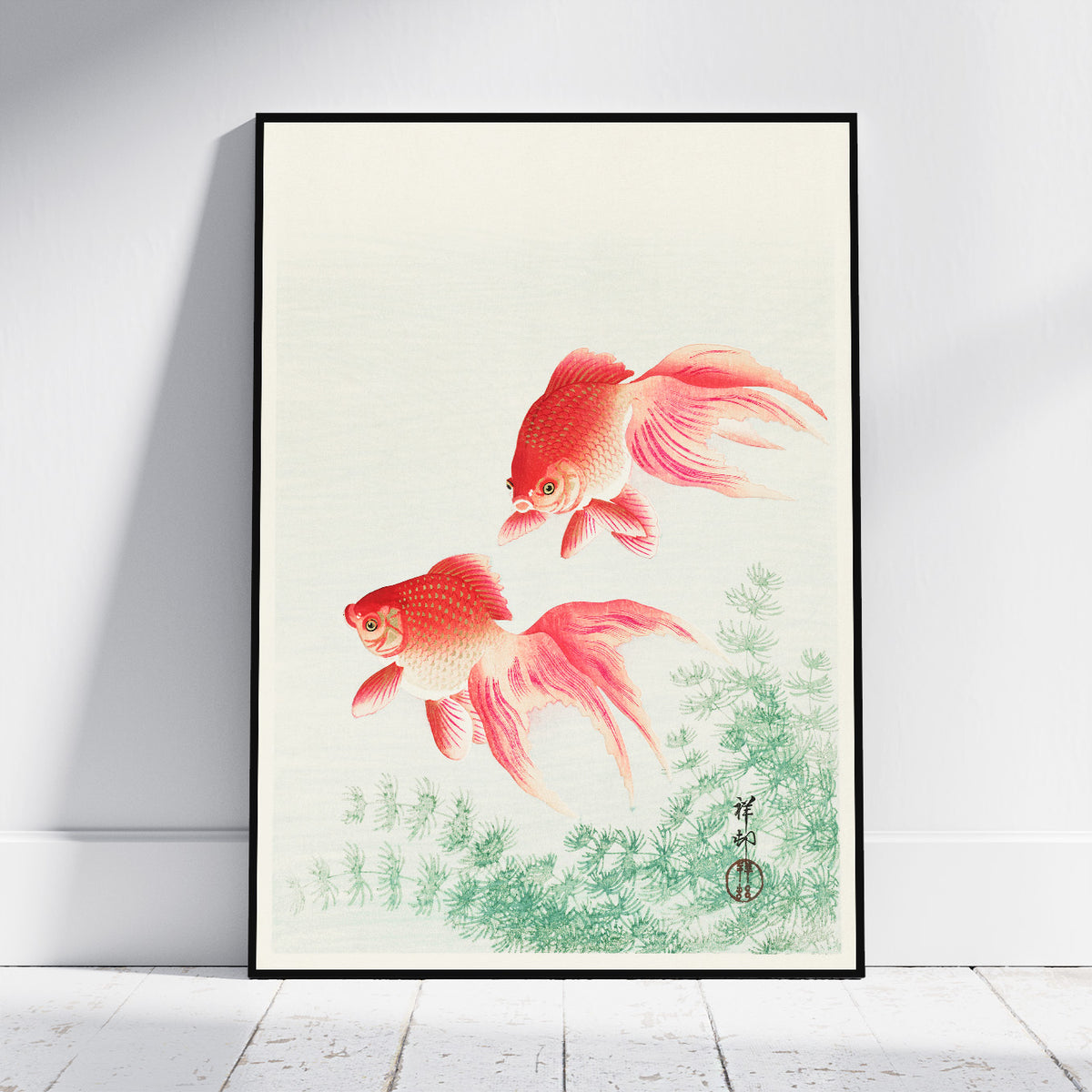 A framed art print featuring two red and orange goldfish swimming above green aquatic plants, displayed against a white wall on a light wood floor.