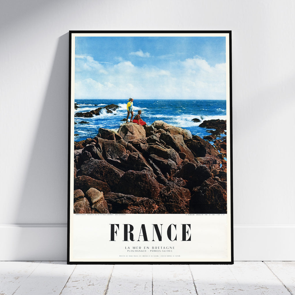 A framed vintage-style travel poster leaning against a white wall on a light wood floor. The poster shows a rocky coastal scene with blue ocean and sky, and the word FRANCE printed at the bottom.