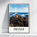 A framed vintage-style travel poster leaning against a white wall on a light wood floor. The poster shows a rocky coastal scene with blue ocean and sky, and the word FRANCE printed at the bottom.
