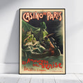 A framed vintage-style poster leaning against a white wall on a light floor. The poster reads "CASINO DE PARIS" and "Les Amoureux de Venise Ballet en 4 tableaux" with illustrated figures in a dark scene and red lettering.