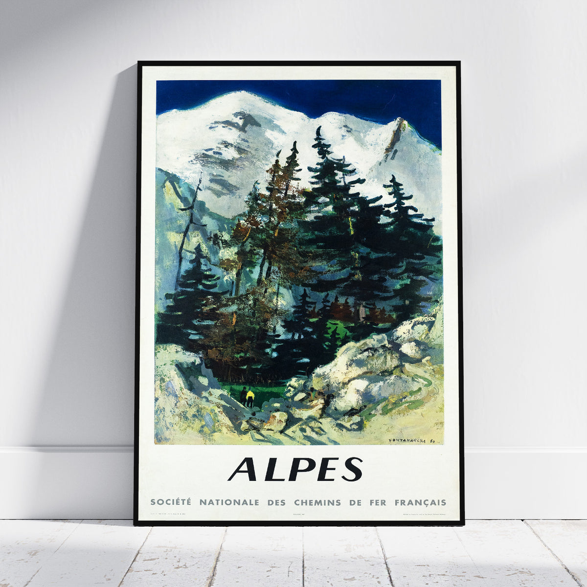 A framed vintage-style travel poster of the Alps, leaning against a white wall on a light wood floor. The poster features a mountain landscape with snow-capped peaks, dark pine trees, and rocky foreground, with the word 'ALPES' and French text at the bottom.
