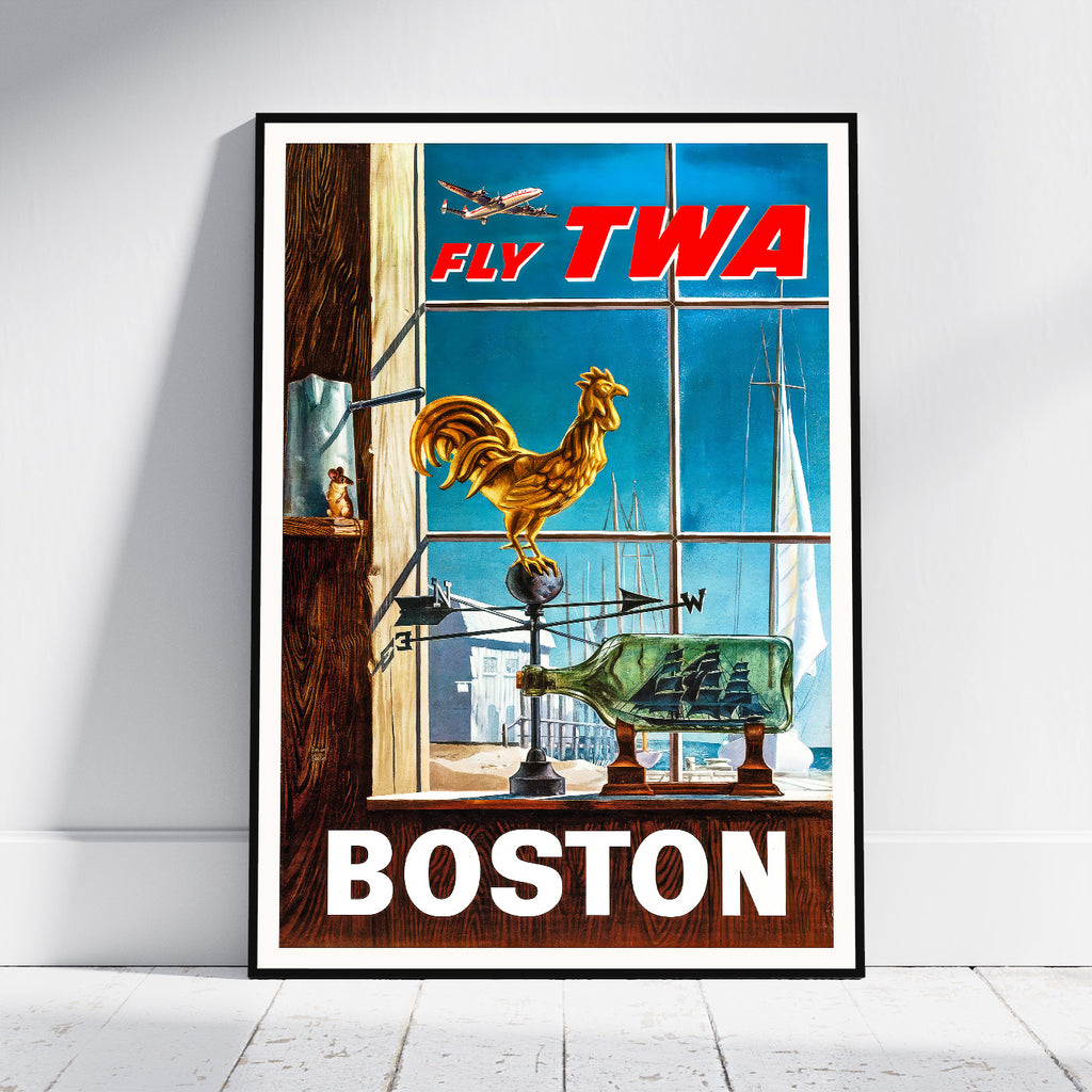 A framed vintage-style travel poster leaning against a white wall on a light floor. The poster shows a Boston scene with large red 'FLY TWA' text at the top, a golden rooster on a weather vane, sailboats and an airplane in the background, and 'BOSTON' in large white letters at the bottom.