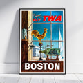 A framed vintage-style travel poster leaning against a white wall on a light floor. The poster shows a Boston scene with large red 'FLY TWA' text at the top, a golden rooster on a weather vane, sailboats and an airplane in the background, and 'BOSTON' in large white letters at the bottom.