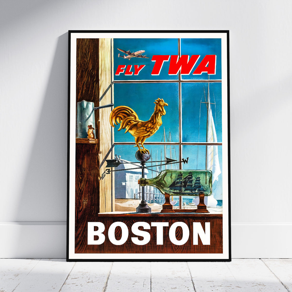 A framed vintage-style travel poster leaning against a white wall on a light floor. The poster shows a Boston scene with large red 'FLY TWA' text at the top, a golden rooster on a weather vane, sailboats and an airplane in the background, and 'BOSTON' in large white letters at the bottom.