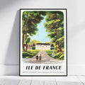 A framed vintage travel poster of Ile de France, leaning against a white wall on a light wood floor. The poster features a retro illustration of a grand chateau, a pathway with two people walking a dog, and lush green trees under a light blue sky.