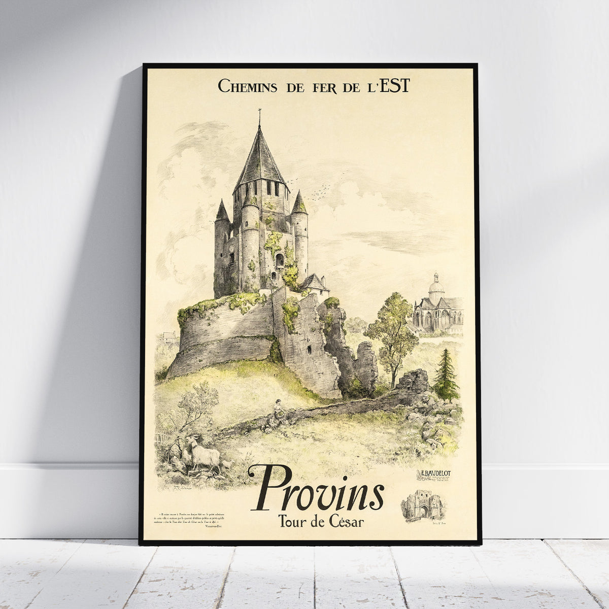 A framed vintage travel poster leaning against a white wall on a light wood floor. The poster features an illustration of a stone castle and historic town with French text reading 'CHEMINS DE FER DE L'EST' and 'Provins Tour de César'.