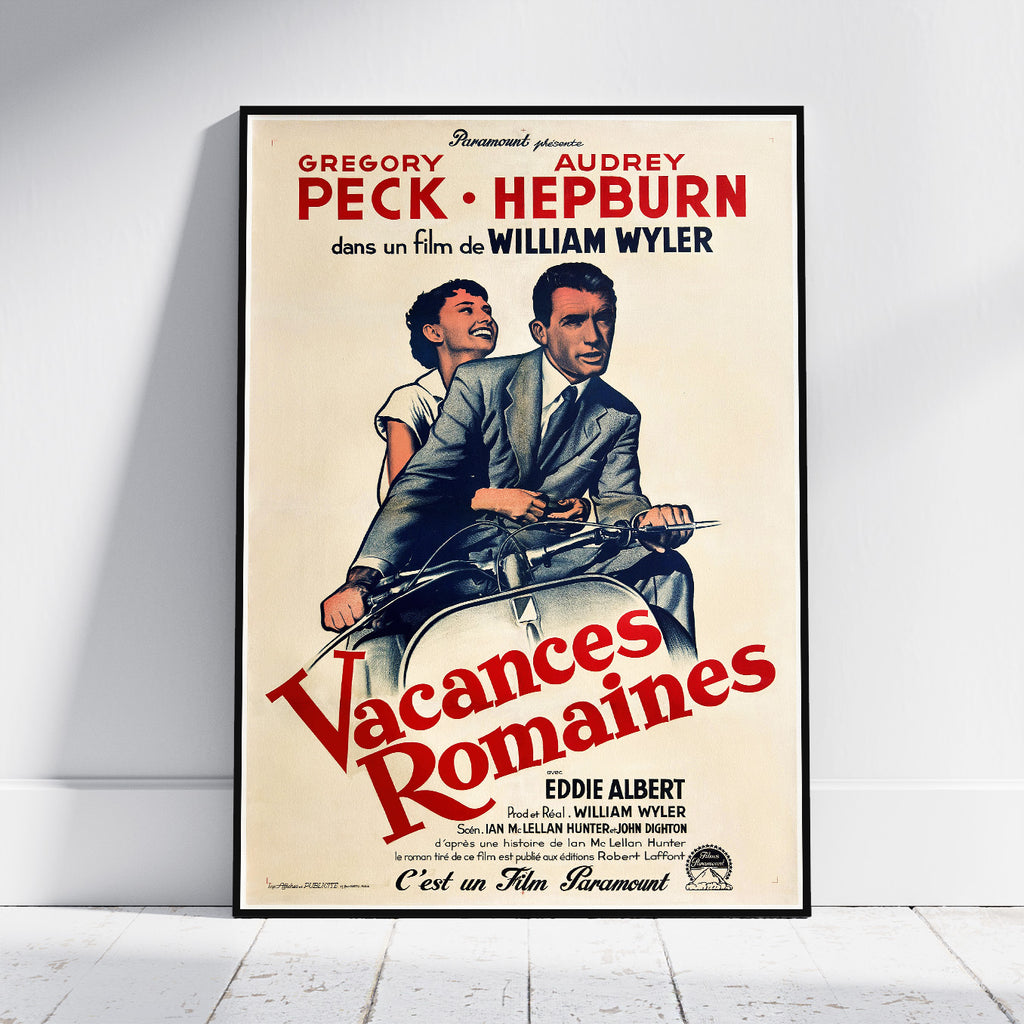 A framed vintage movie poster for Gregory Peck and Audrey Hepburn's film 'Vacances Romaines' by Paramount, leaning against a white wall on a light wood floor.