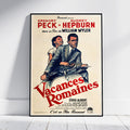 A framed vintage movie poster for Gregory Peck and Audrey Hepburn's film 'Vacances Romaines' by Paramount, leaning against a white wall on a light wood floor.