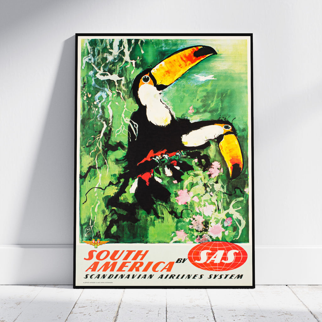 A framed vintage-style travel poster leaning against a white wall on a light wood floor. The artwork shows two toucans in a green jungle scene with flowers, and the text 'SOUTH AMERICA BY SAS' at the bottom.