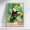 A framed vintage-style travel poster leaning against a white wall on a light wood floor. The artwork shows two toucans in a green jungle scene with flowers, and the text 'SOUTH AMERICA BY SAS' at the bottom.