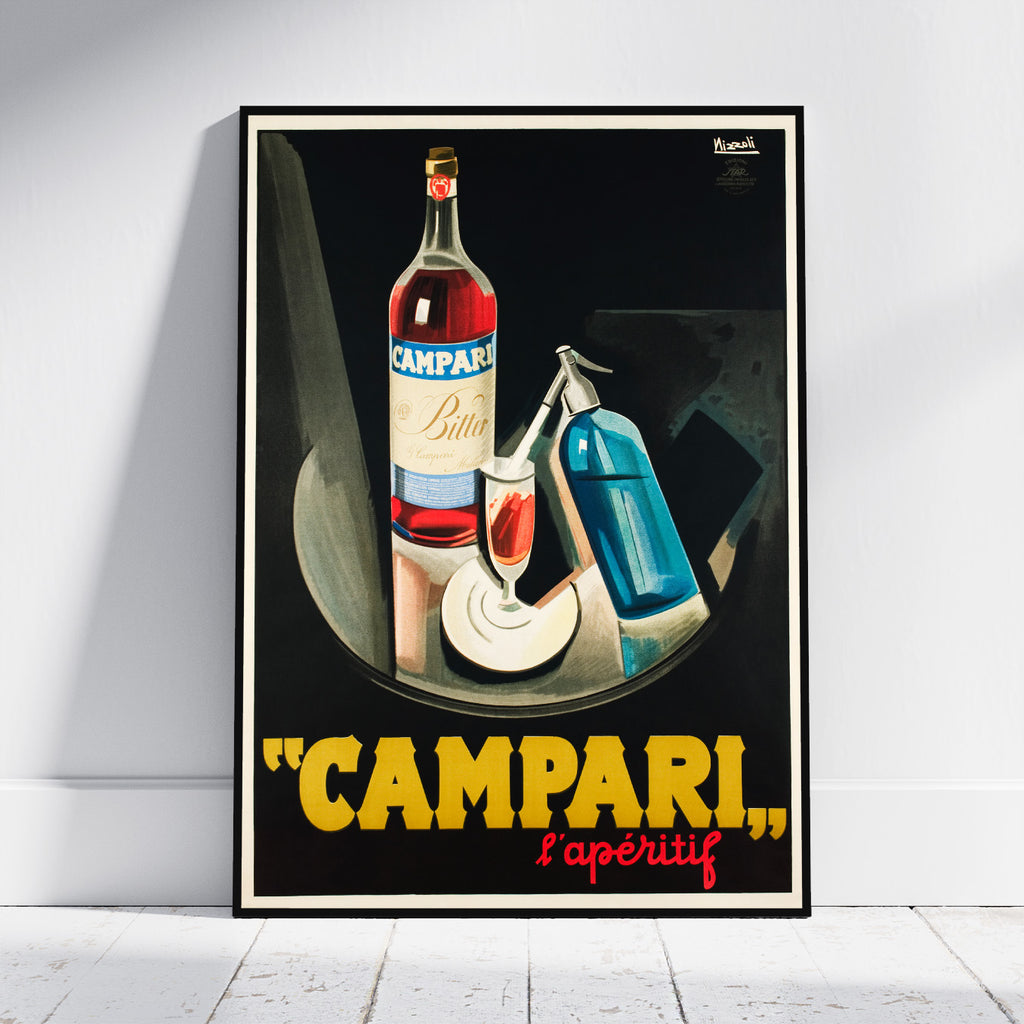 A framed vintage-style poster leaning against a white wall on a light floor. The poster shows a Campari bottle, a filled glass, and a blue bottle on a dark background, with large yellow text reading "CAMPARI" and smaller red text reading "l'apéritif" at the bottom.