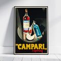 A framed vintage-style poster leaning against a white wall on a light floor. The poster shows a Campari bottle, a filled glass, and a blue bottle on a dark background, with large yellow text reading "CAMPARI" and smaller red text reading "l'apéritif" at the bottom.