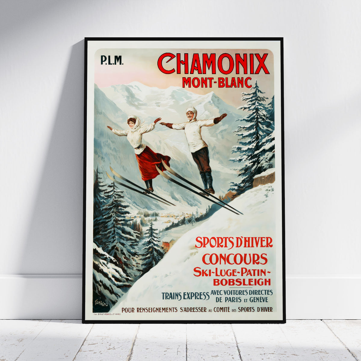 A framed vintage-style Chamonix Mont-Blanc poster leaning against a white wall on a light floor. The poster shows two skiers in a snowy mountain landscape with red and black text, and a black frame around the print.
