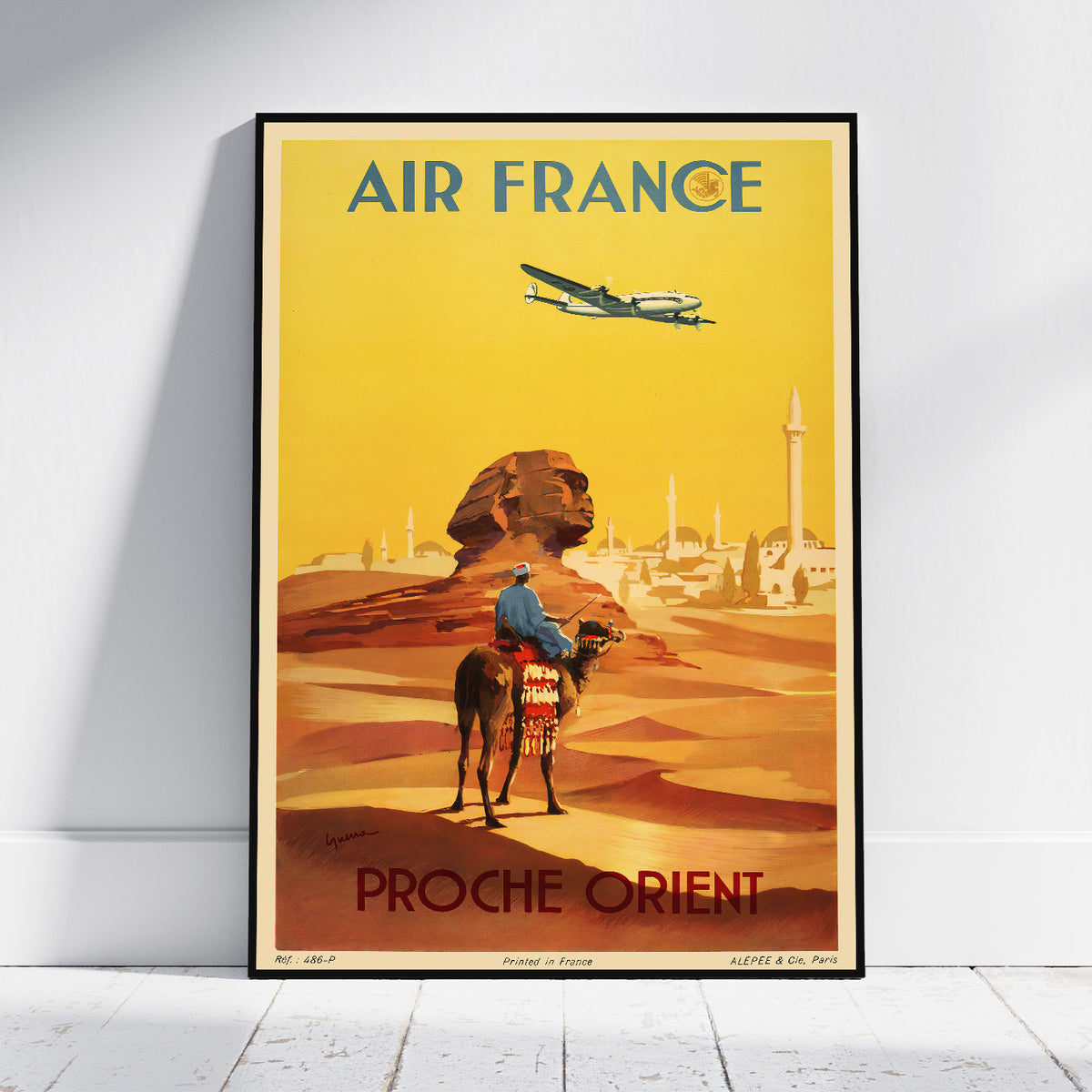A framed vintage-style Air France travel poster leaning against a white wall on a light floor. The poster shows a yellow desert scene with a camel rider, sand dunes, a lion statue, and an airplane, with the text 'AIR FRANCE' at the top and 'PROCHE ORIENT' at the bottom.