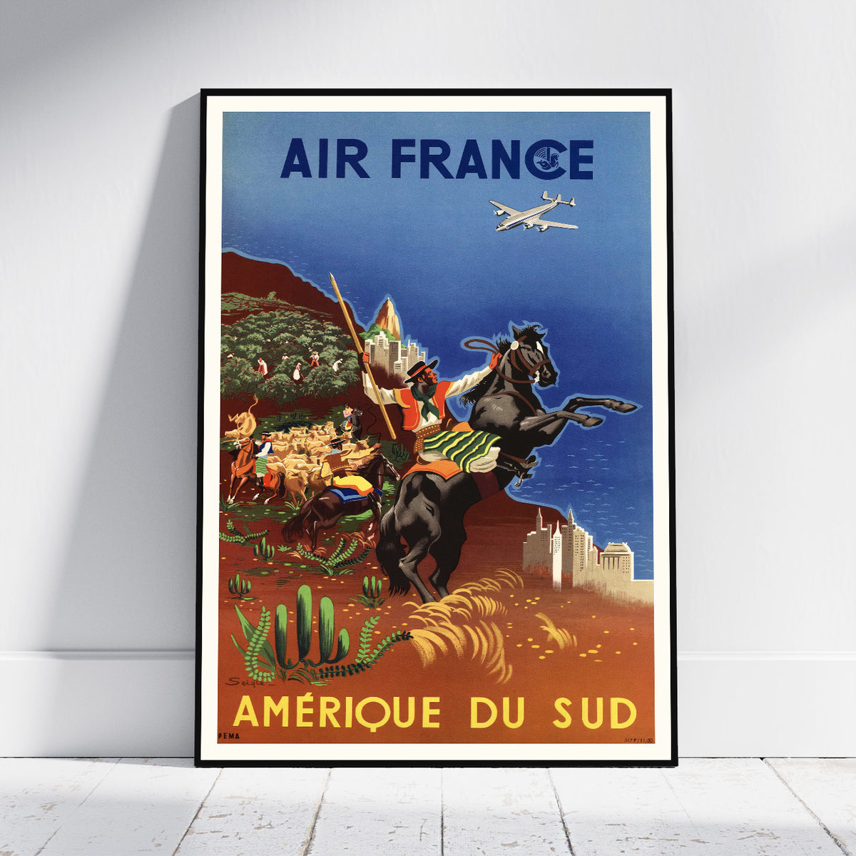 A framed vintage-style travel poster leaning against a white wall on a light floor. The poster reads "AIR FRANCE" at the top and "AMÉRIQUE DU SUD" at the bottom, showing an illustrated scene with a rider on horseback, desert landscape, cacti, and an airplane.