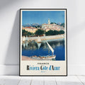 A framed vintage-style travel poster of Riviera Còte d'Azur, France, leaning against a white wall on a light wood floor. The poster features a scenic illustration of a coastal town with sailboats on the water under a blue sky, with 'FRANCE Riviera Còte d'Azur' printed at the bottom.