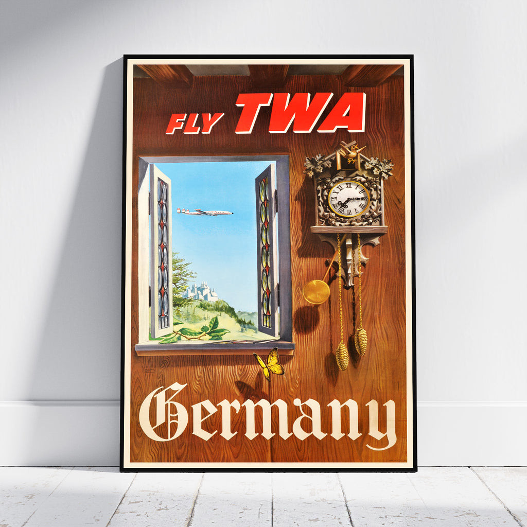 A framed vintage travel poster leaning against a white wall on a light floor. The poster reads 'FLY TWA' at the top and 'Germany' at the bottom, with a wooden door background, a cuckoo clock, and an open window showing an airplane and green landscape.
