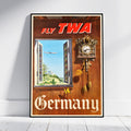 A framed vintage travel poster leaning against a white wall on a light floor. The poster reads 'FLY TWA' at the top and 'Germany' at the bottom, with a wooden door background, a cuckoo clock, and an open window showing an airplane and green landscape.