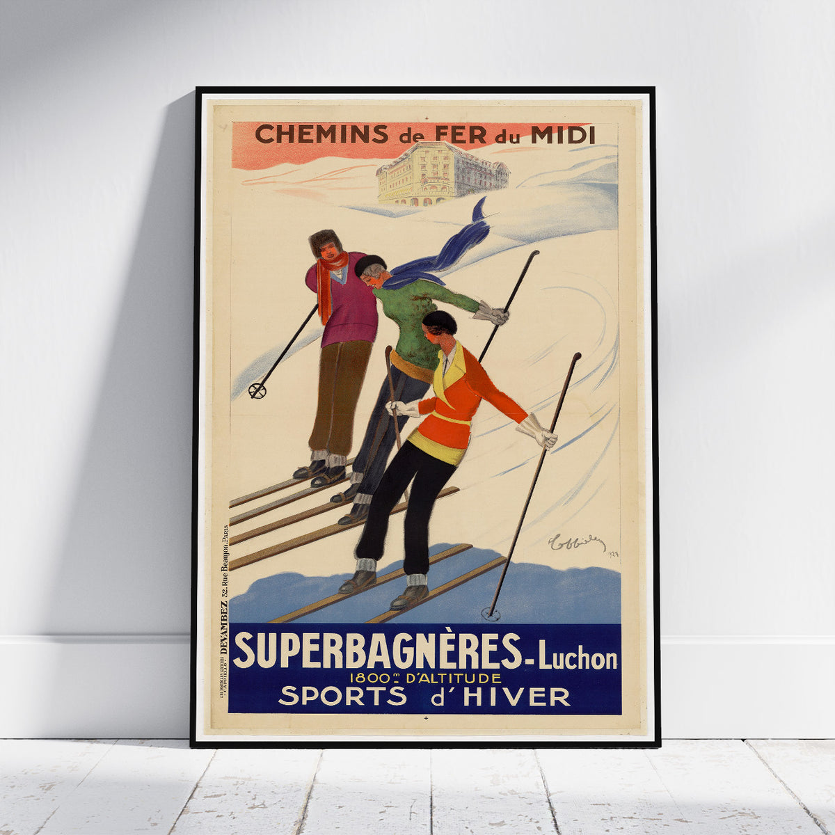 A framed vintage travel poster for French railways advertising 'Superbagnères - Luchon' ski resort, leaning against a white wall on a light wood floor. The poster features three skiers on a snowy slope with a cream background and bold blue and orange text.