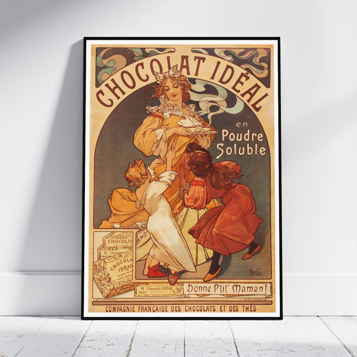 A framed vintage-style chocolate advertisement poster leaning against a white wall on a light floor. The poster features an Art Nouveau illustration with the text 'CHOCOLAT IDEAL en Poudre Soluble' and depicts a woman, a child, and a dog in warm cream, rust, and brown tones.