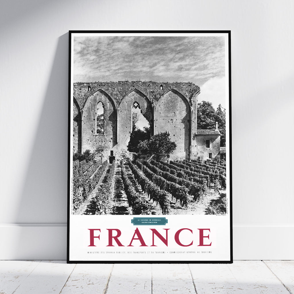 A framed black and white travel poster of a French vineyard landscape leaning against a white wall on a light wooden floor. The poster features a black and white photograph of a stone cloister and vineyard rows, with 'FRANCE' printed in large red letters at the bottom.