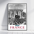 A framed black and white travel poster of a French vineyard landscape leaning against a white wall on a light wooden floor. The poster features a black and white photograph of a stone cloister and vineyard rows, with 'FRANCE' printed in large red letters at the bottom.