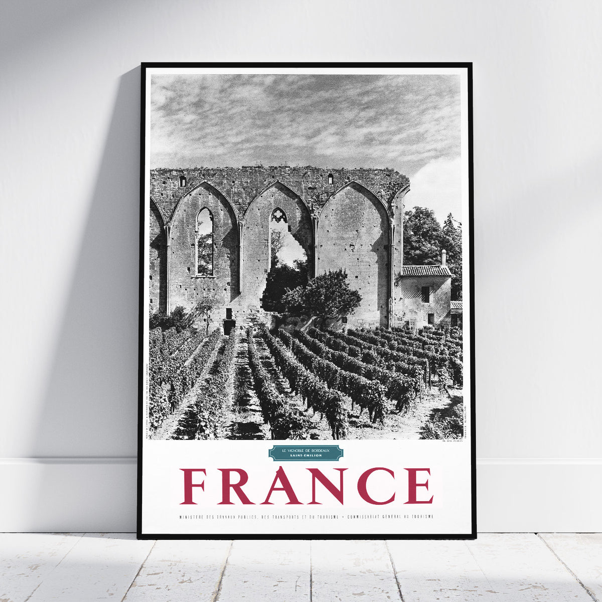 A framed black and white travel poster of a French vineyard landscape leaning against a white wall on a light wooden floor. The poster features a black and white photograph of a stone cloister and vineyard rows, with 'FRANCE' printed in large red letters at the bottom.