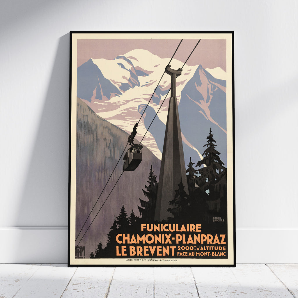 A framed vintage-style travel poster leaning against a white wall on a light floor. The poster shows snow-covered mountains, evergreen trees, and a cable car system with French text reading 'Funiculaire Chamonix-Planpraz Le Brevent'.