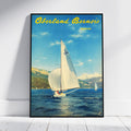 A framed vintage-style travel poster leaning against a white wall on a light wood floor. The poster shows a sailboat on blue water with mountains and green cliffs in the background, and the text 'Oberland Bernois' in yellow at the top.