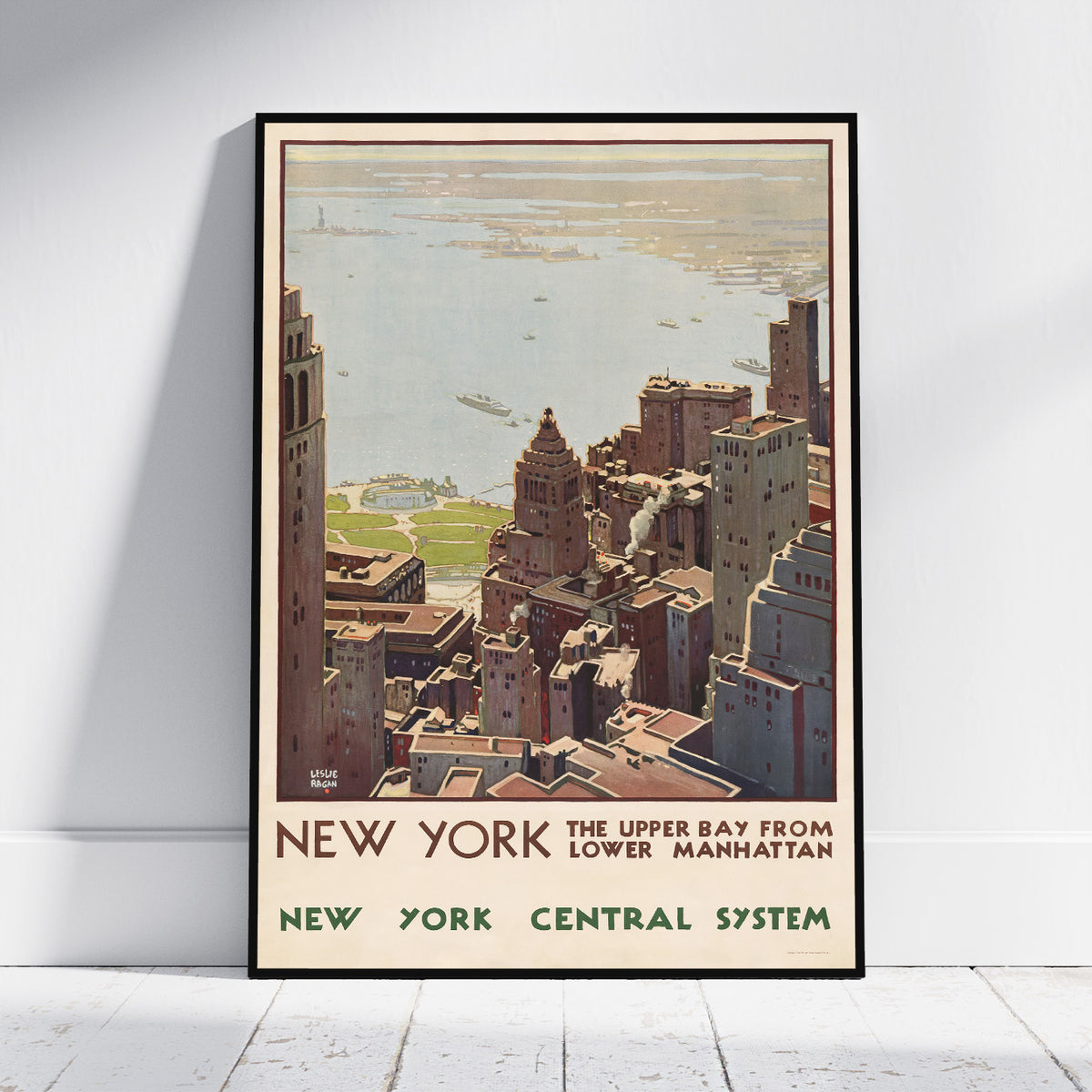 A framed vintage-style New York travel poster leaning against a white wall on a light floor. The poster shows an aerial view of the city skyline and water with text reading 'NEW YORK THE UPPER BAY FROM LOWER MANHATTAN' and 'NEW YORK CENTRAL SYSTEM'.