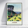 A framed vintage travel poster of Jura, France, leaning against a white wall on a light wood floor. The poster features a colorful impressionist landscape painting with a church, green hills, and a large rock formation under a pale blue sky, with 'JURA' printed at the bottom.