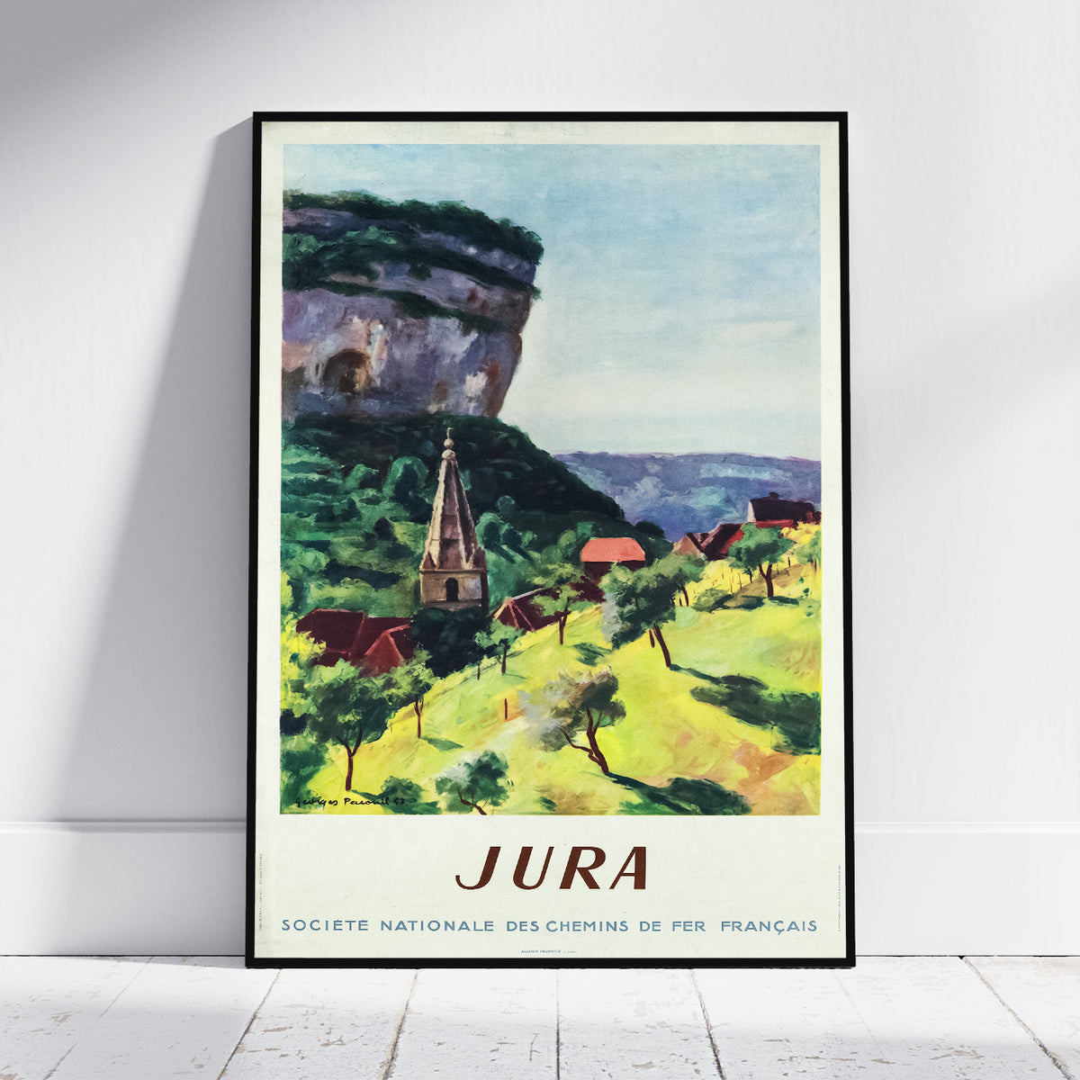 A framed vintage travel poster of Jura, France, leaning against a white wall on a light wood floor. The poster features a colorful impressionist landscape painting with a church, green hills, and a large rock formation under a pale blue sky, with 'JURA' printed at the bottom.