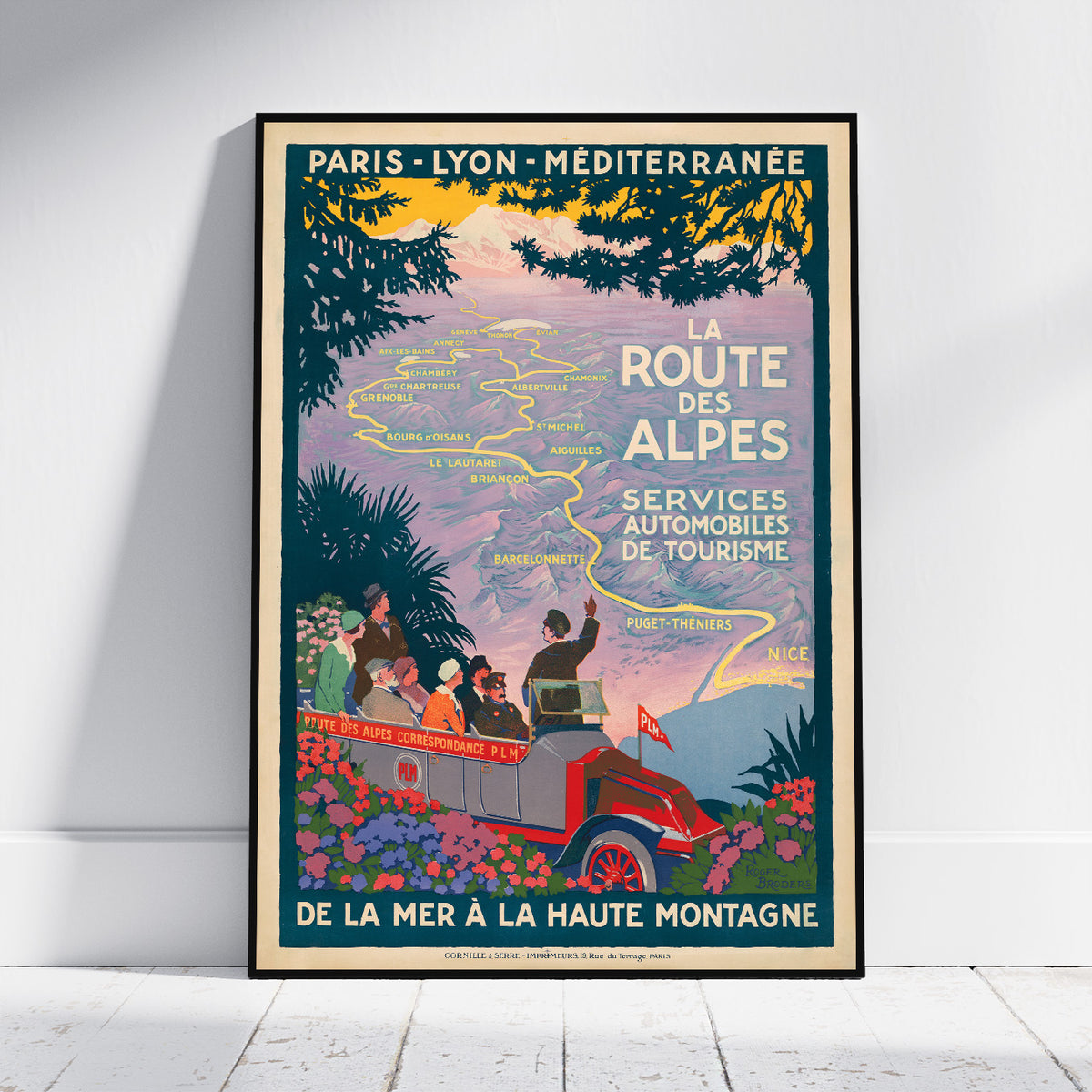 A vintage-style travel poster for the French Alps, leaning against a white wall on a light wood floor. The poster features a retro car with passengers, a map of the region, and text reading 'LA ROUTE DES ALPES' and 'PARIS - LYON - MÉDITERRANÉE'.