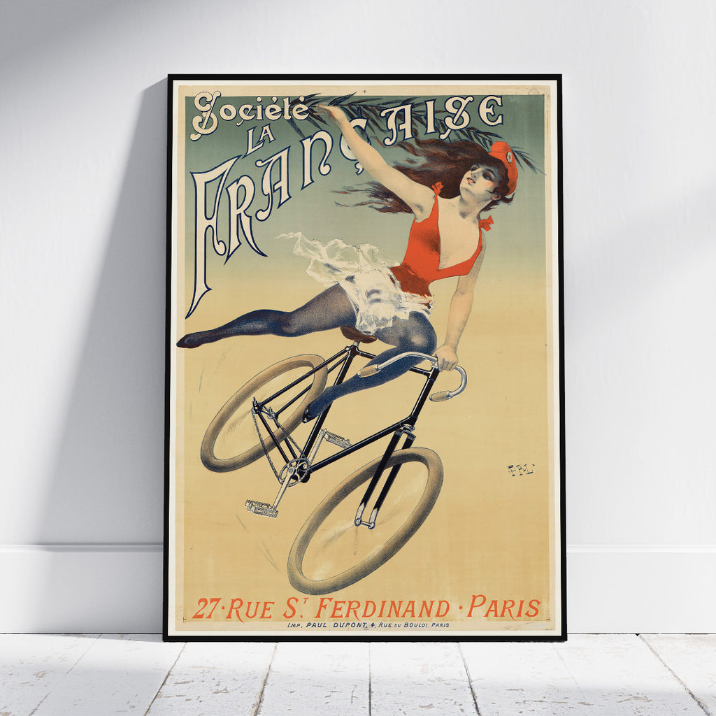 A framed vintage-style poster leaning against a white wall on a light floor. The poster shows an illustrated woman riding a bicycle with large decorative text reading 'Société La Française' and additional text at the bottom.