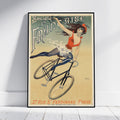 A framed vintage-style poster leaning against a white wall on a light floor. The poster shows an illustrated woman riding a bicycle with large decorative text reading 'Société La Française' and additional text at the bottom.