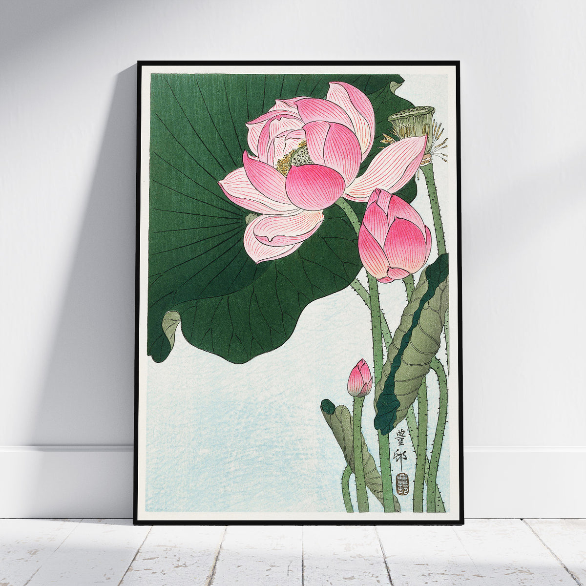 A framed art print featuring a traditional Japanese-style illustration of pink lotus flowers and large green lotus leaves, leaning against a white wall on a light wood floor.