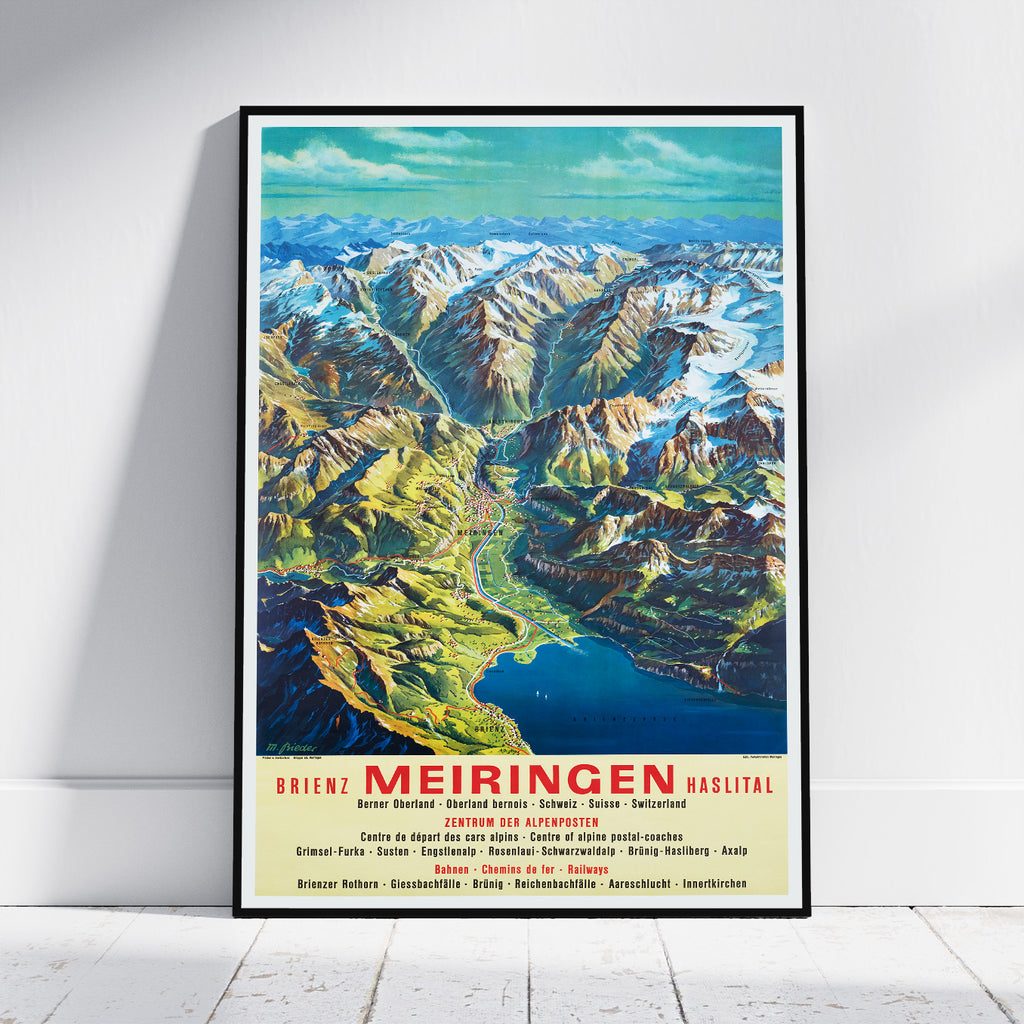A framed vintage-style travel poster leaning against a white wall on a light floor. The poster shows a colorful mountain landscape with a valley, blue lake, and bold red text reading 'BRIENZ MEIRINGEN HAGLITAL' at the bottom.