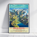 A framed vintage-style travel poster leaning against a white wall on a light floor. The poster shows a colorful mountain landscape with a valley, blue lake, and bold red text reading 'BRIENZ MEIRINGEN HAGLITAL' at the bottom.