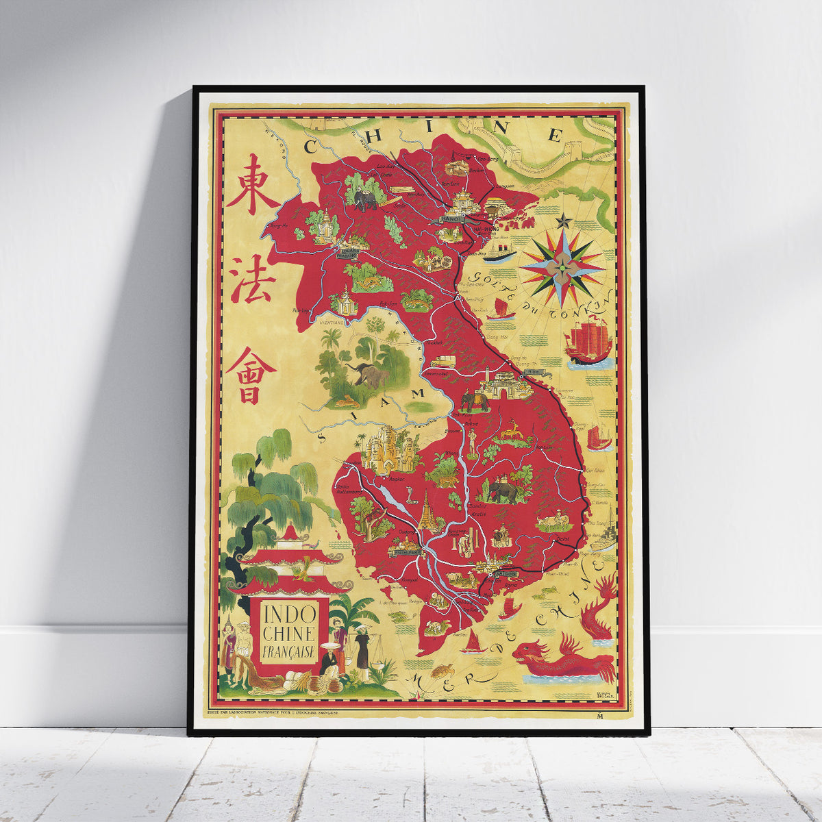 A framed vintage-style map poster of Indochina, leaning against a white wall on a light wood floor. The map features a warm beige background with red highlighted regions, green illustrations, and decorative cartographic details.