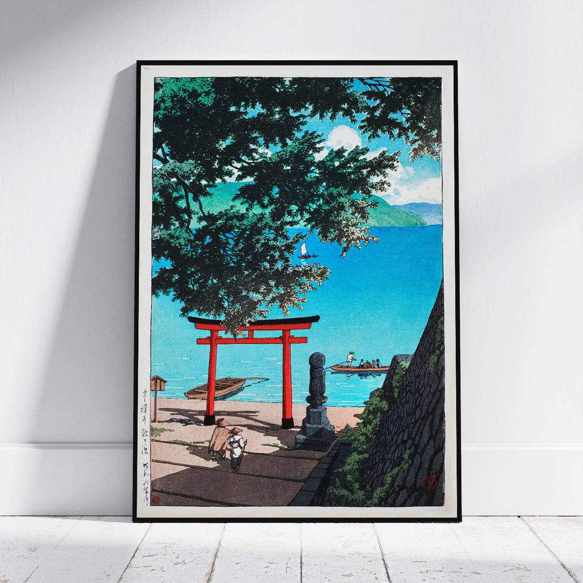 A framed art print featuring a Japanese-style coastal scene with a bright red torii gate, blue ocean, trees, and a stone wall, leaning against a white wall on a light floor.