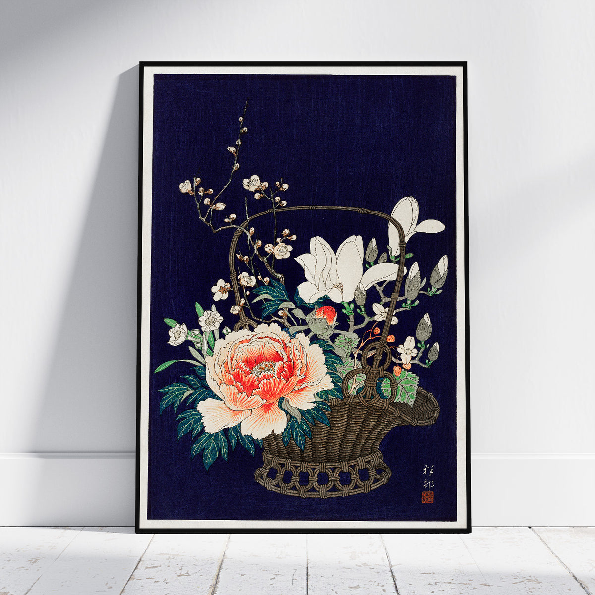 A framed art print featuring a Japanese-style floral illustration of a wicker basket filled with white lilies, a large peach and pink peony, and green leaves, set against a dark navy blue background. The print is framed in black and leans against a white wall on a light wood floor.