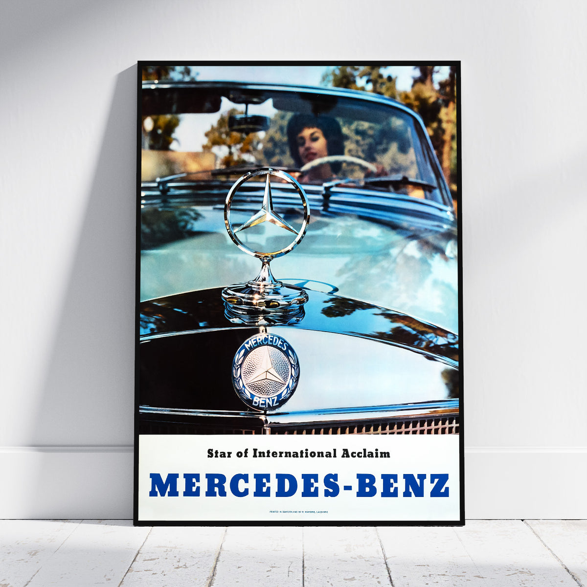 A framed vintage-style Mercedes-Benz poster leaning against a white wall on a light wood floor. The poster shows a close-up of a classic car hood with the Mercedes-Benz star emblem and badge, plus a blurred figure in the windshield and large blue 'MERCEDES-BENZ' text at the bottom.
