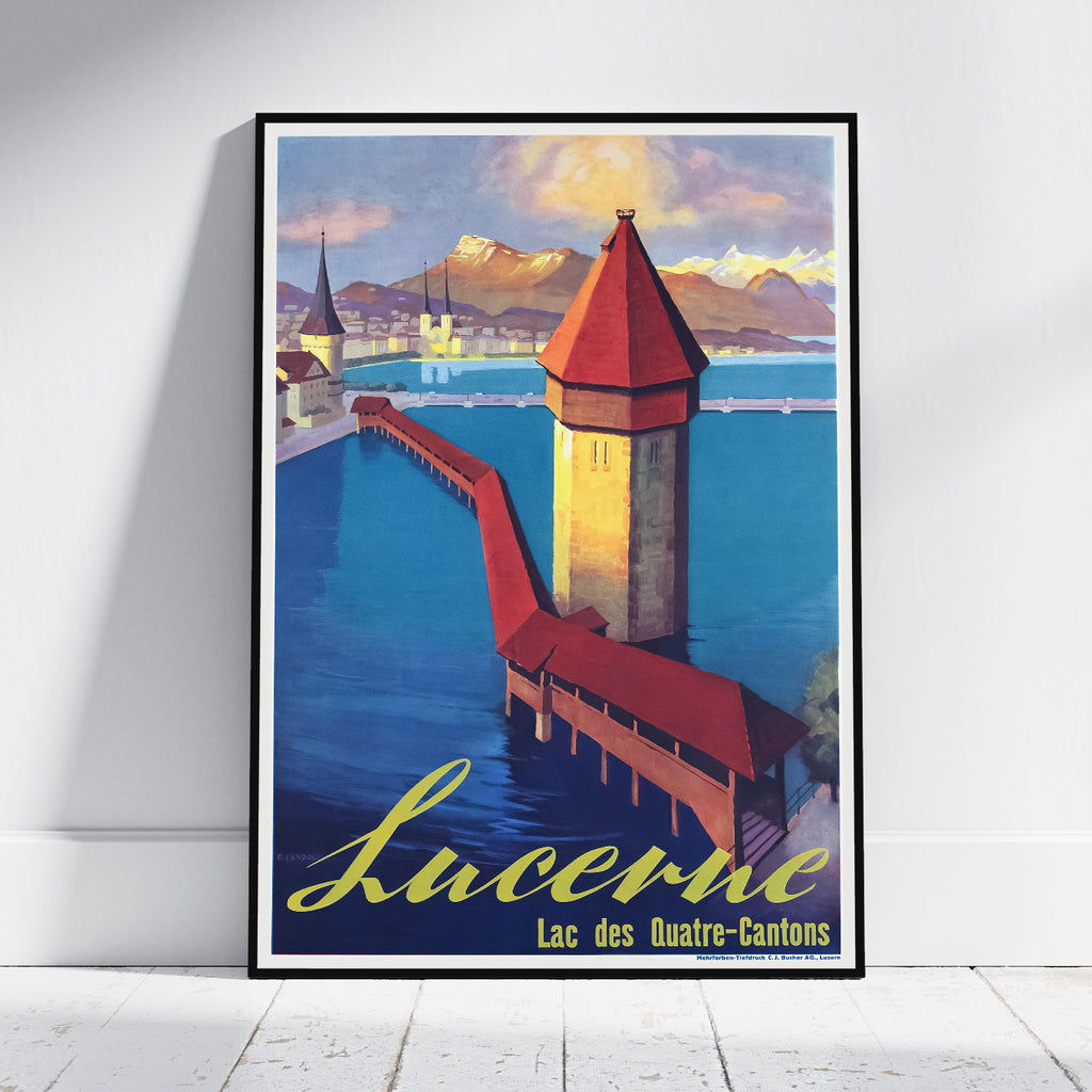 A framed vintage-style travel poster leaning against a white wall on a light wood floor. The poster depicts a scenic view of Lucerne, Switzerland, with the Chapel Bridge, Water Tower, blue lake, mountains, and the word 'Lucerne' in large yellow script at the bottom.