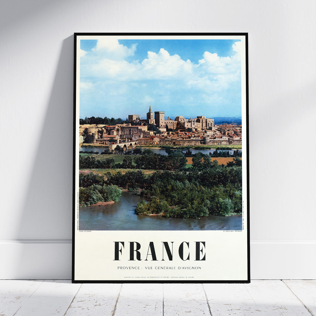 A framed vintage-style travel poster of France, leaning against a white wall on a light wood floor. The poster features a scenic view of a historic French city by a river under a blue sky, with 'FRANCE' printed in bold black letters at the bottom.
